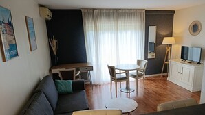 Una sala de estar moderna con un sofá, una mesa de comedor con sillas, un televisor sobre un mueble bajo y un ventanal con cortinas translúcidas.