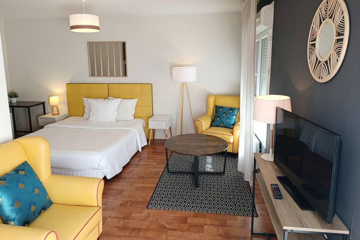 A hotel room with a bed, two yellow armchairs, a TV, and a small table.