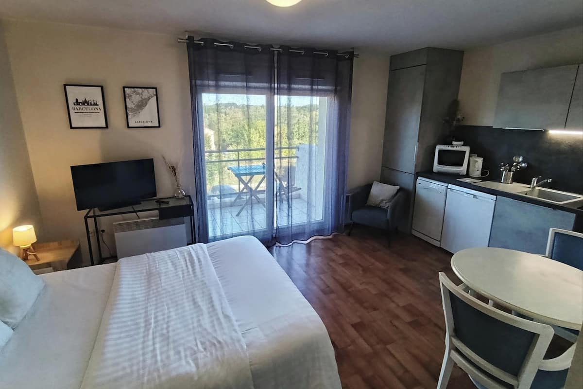 A hotel room with a bed, a TV, a balcony with a table and chairs, a kitchenette, and framed pictures on the wall.