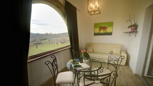 A room with a round glass table, two chairs, a sofa, a painting of a cow, and a view of a green landscape through a large window. - Pietriccio Rosso (Manciano)