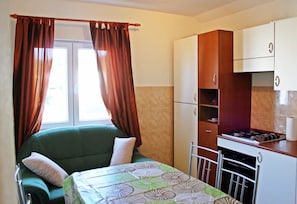 Una habitación compacta con un sofá verde, una pequeña mesa de comedor, una kitchenette con estufa y una ventana con cortinas.