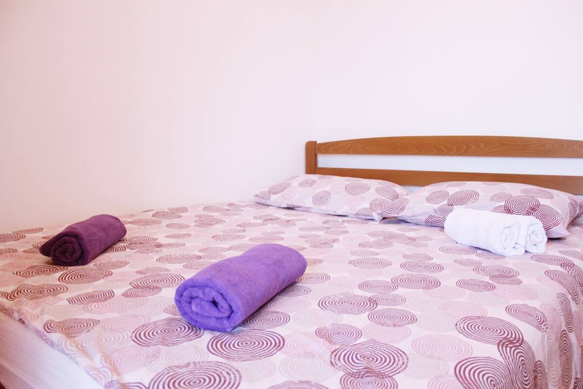 Uma cama com colcha estampada, duas toalhas enroladas e cabeceira de madeira.