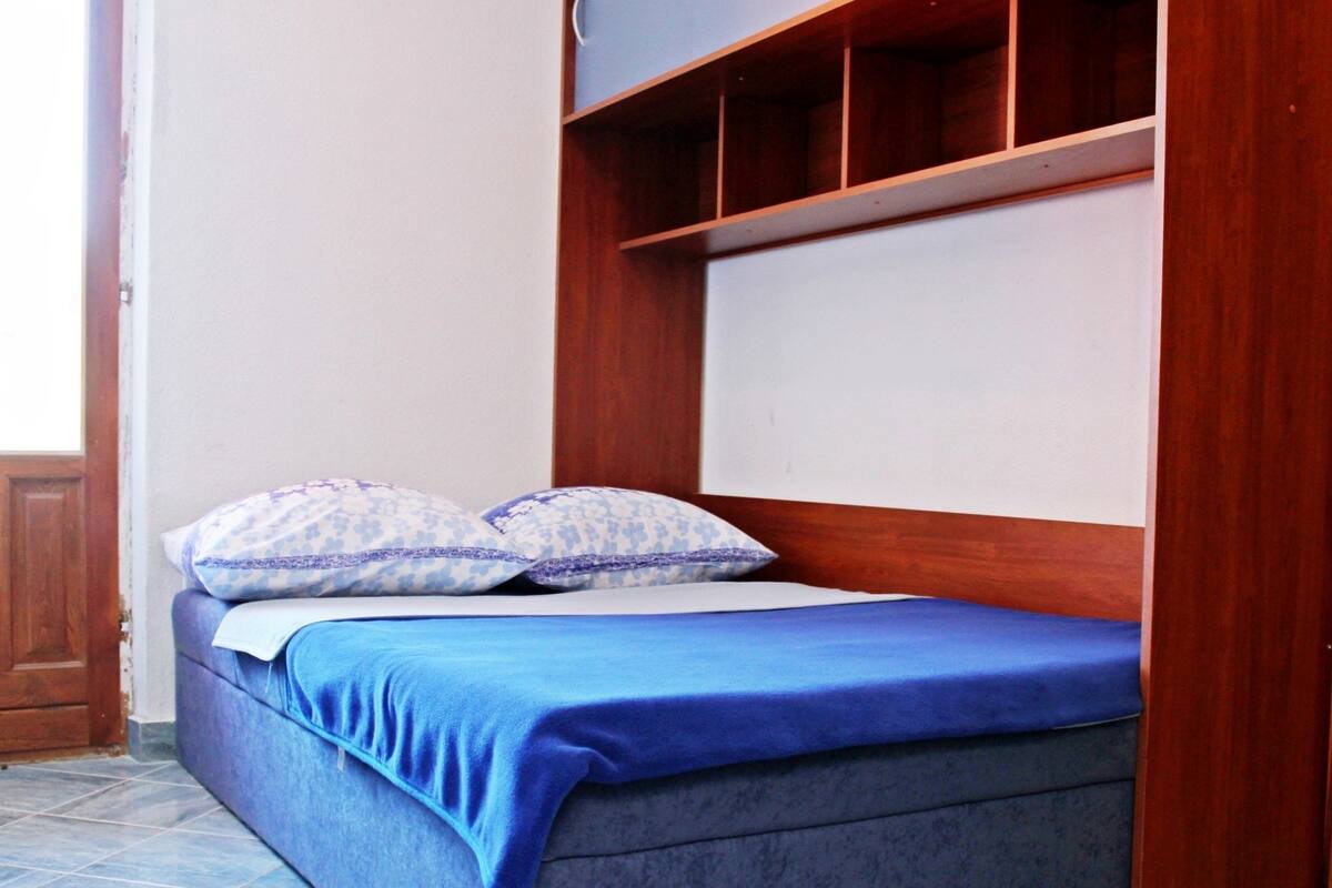Cama de solteiro com colchão azul e roupa de cama combinando, duas almofadas estampadas e cabeceira de madeira.