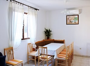 Un comedor con una mesa preparada para cuatro personas, rodeada de sillas de madera con cojines a rayas. Hay una planta en maceta en el alféizar de la ventana, un cuadro en la pared y un equipo de aire acondicionado sobre la mesa.