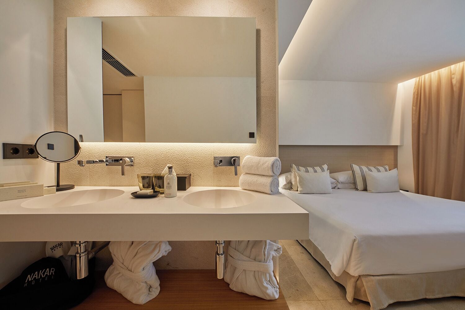 A hotel room with a double sink, a large mirror, a bed with white bedding, and a robe hanging on the floor.