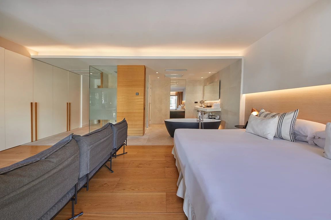 A modern hotel room with a large bed, a desk, a chair, a mirror, and a bathroom visible through a glass partition.