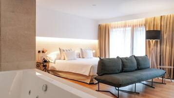 A modern hotel room with a large bed, a sofa, a bathtub, and a desk with a lamp.