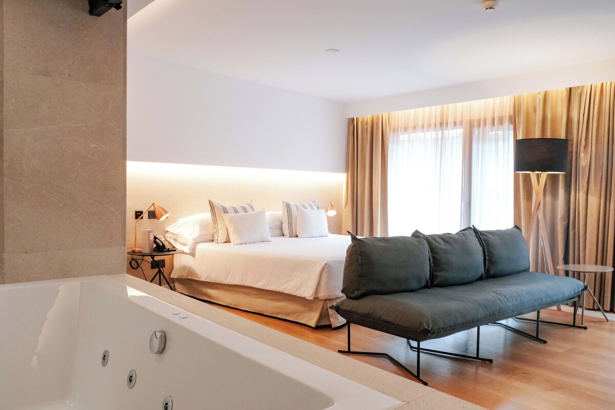 Een hotelkamer met een groot bed, een bank, een badkuip en een klein tafeltje met een lamp.