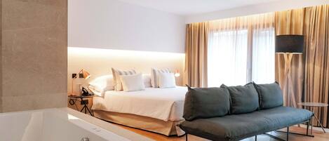 A hotel room with a large bed, a sofa, a bathtub, and a small table with a lamp.