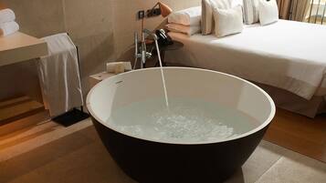 A modern bathroom with a large bathtub, a bedside table with a lamp, and a neatly made bed.