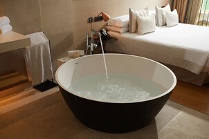 Un baño moderno con una bañera grande, una mesita de noche con lámpara y una cama bien hecha.