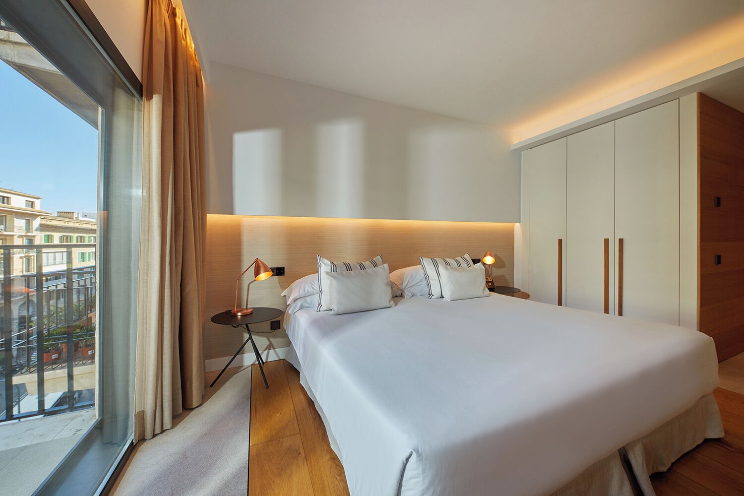 A modern hotel room with a large bed, bedside lamps, and a view of the city through a large window.