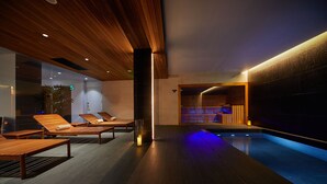 Una piscina interior moderna con tumbonas de madera, zona de sauna y suelo oscuro.