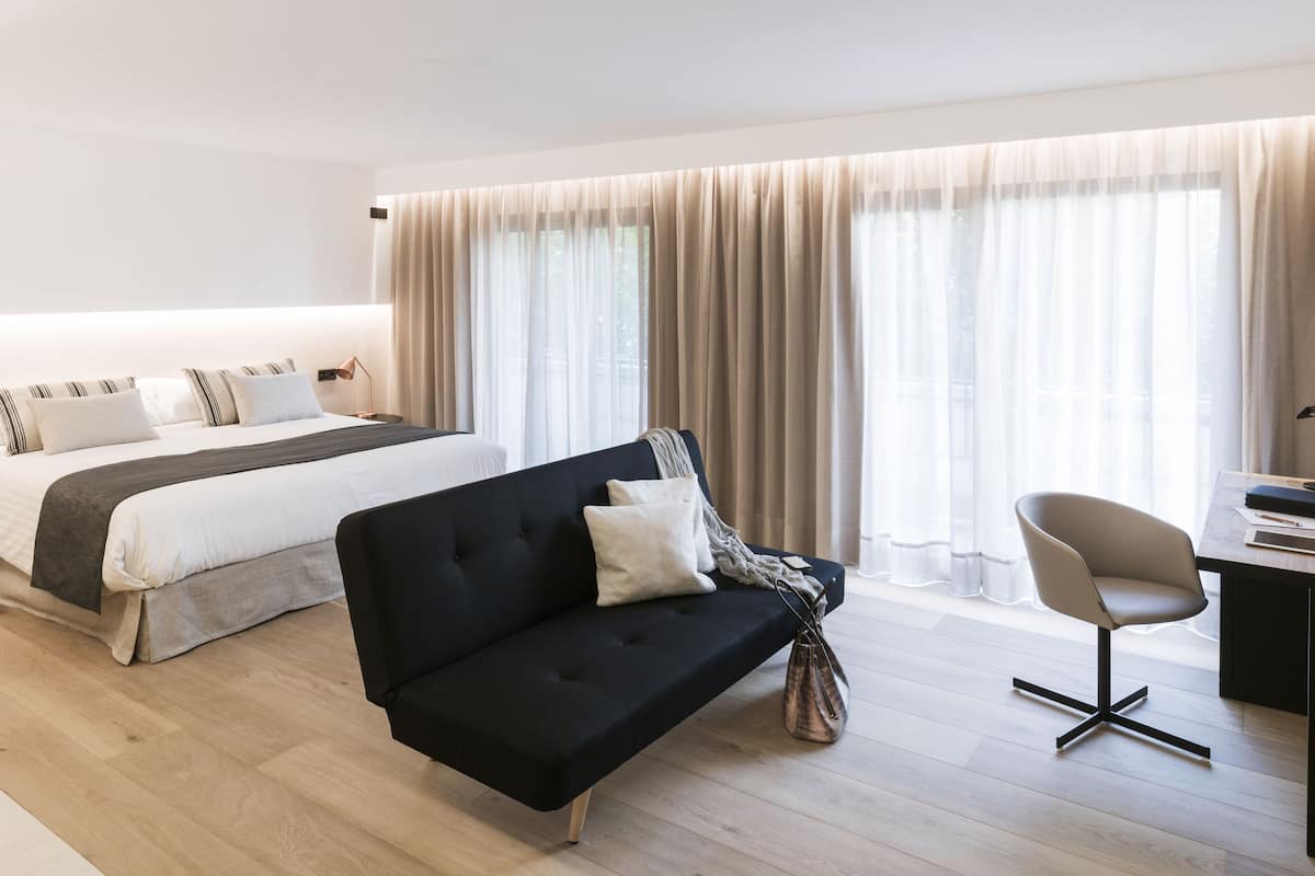Een moderne hotelkamer met een groot bed, een zwarte bank, een bureau met stoel en een groot raam met gordijnen.