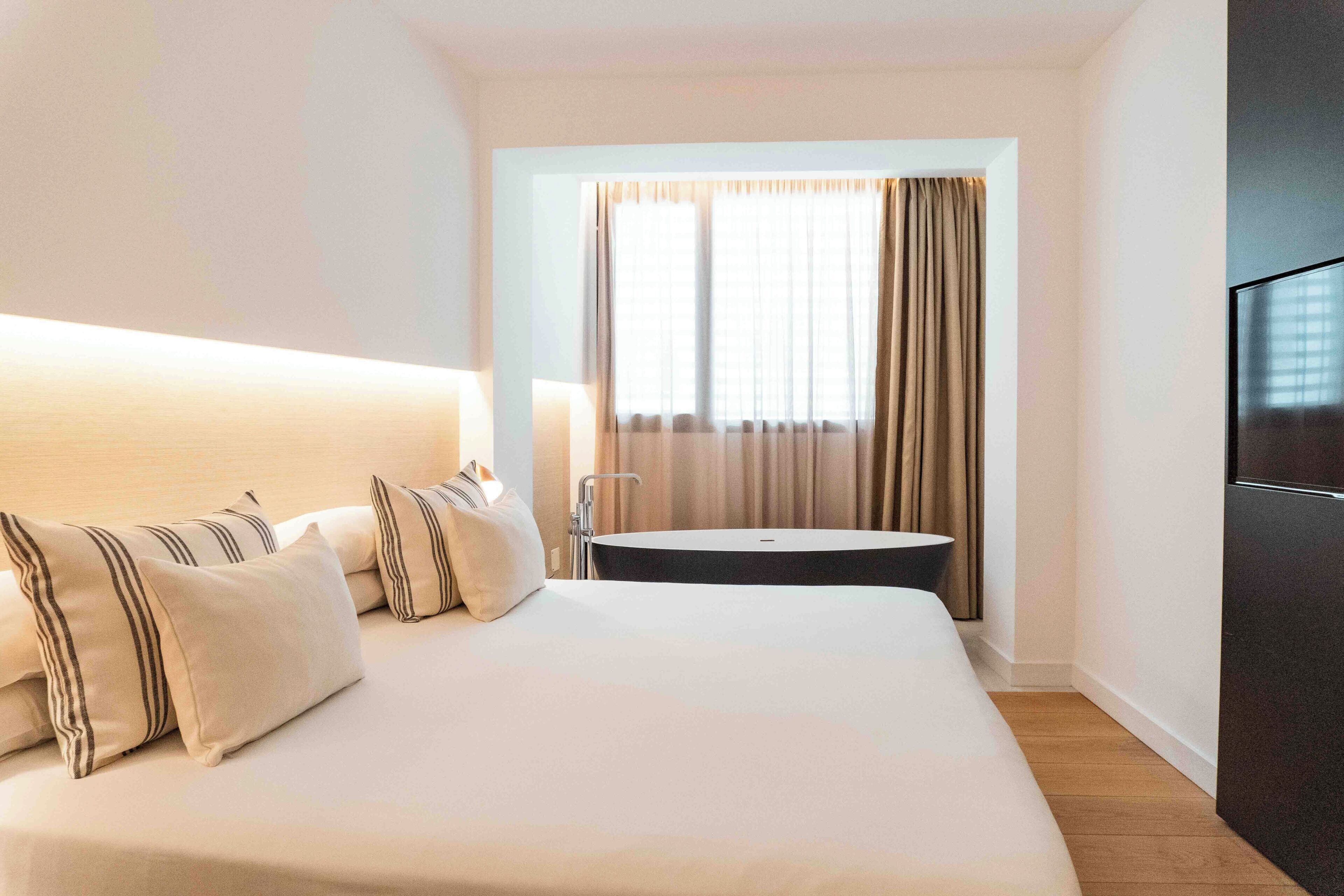 A hotel room with a large bed, a bedside table, a TV, a window with curtains, and a bathroom with a sink and mirror.
