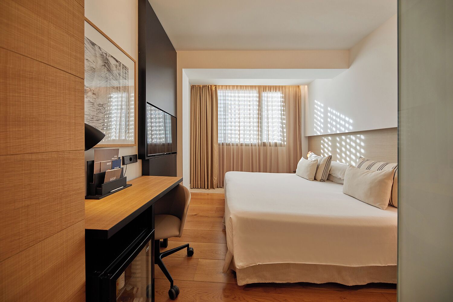 A hotel room with a bed, a desk with a computer, a television, and a window with curtains.