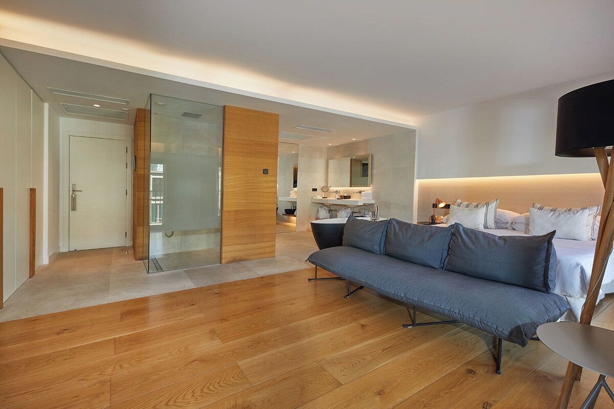 Een moderne hotelkamer met een houten vloer, een grijze bank, een nachtkastje en een grote spiegel.
