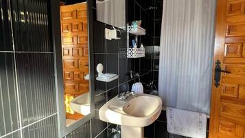 Baño con lavamanos, espejo y zona de ducha. Se aprecia una puerta de madera y una toalla blanca colgada en la pared.