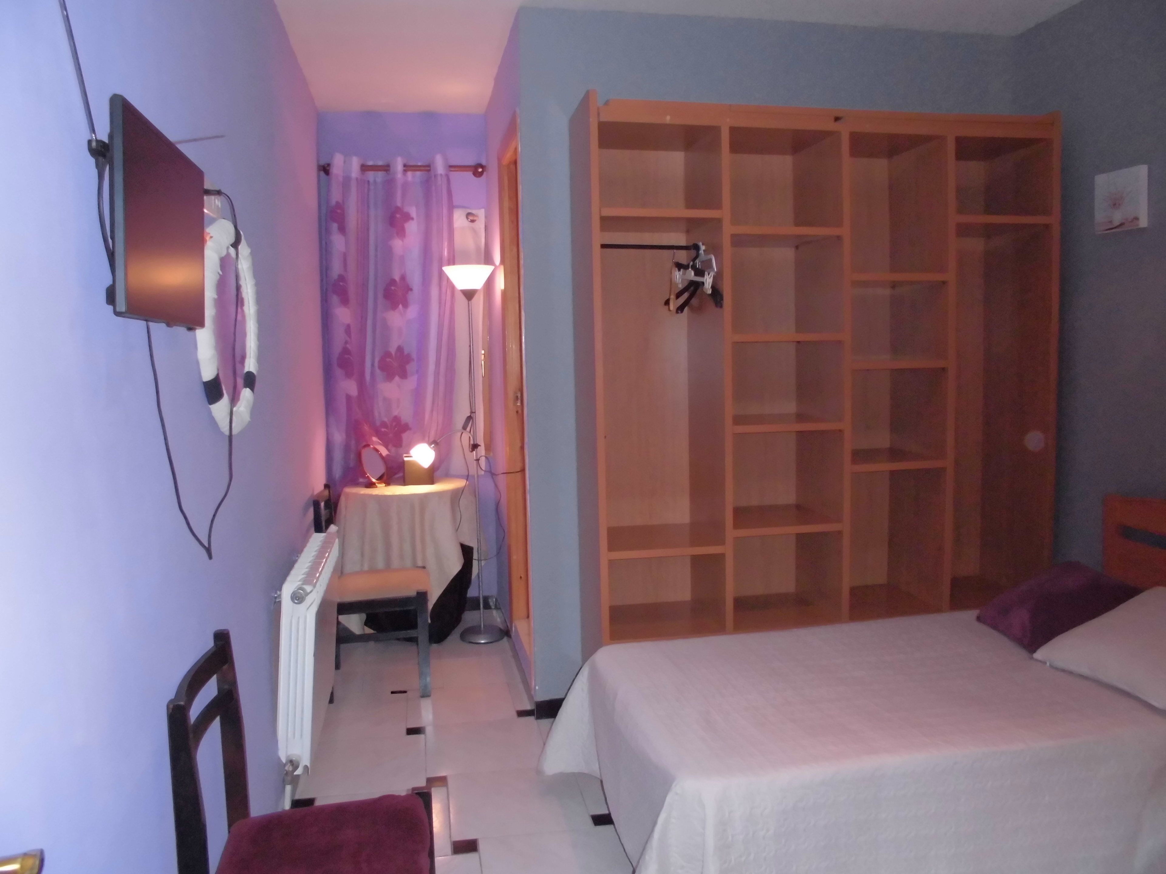 A bedroom with a bed, a wooden wardrobe, a desk with a lamp, and a mirror.