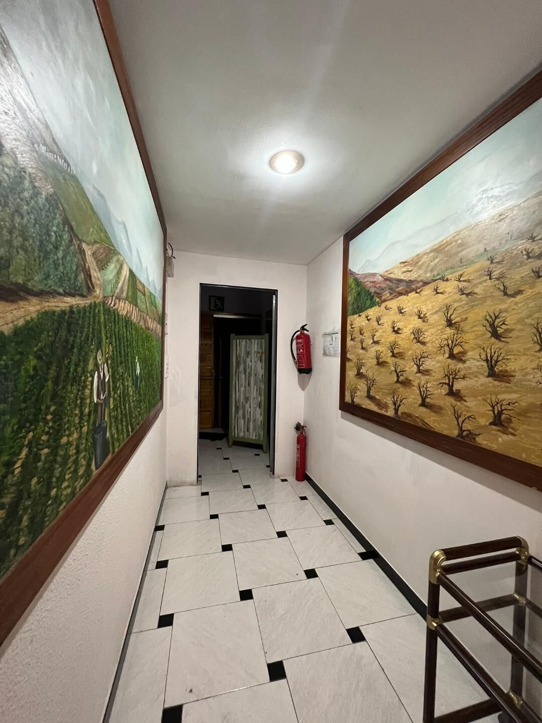 A hallway with tiled flooring, two large landscape murals on the walls, a fire extinguisher mounted on the wall, and a wooden bench on the right.