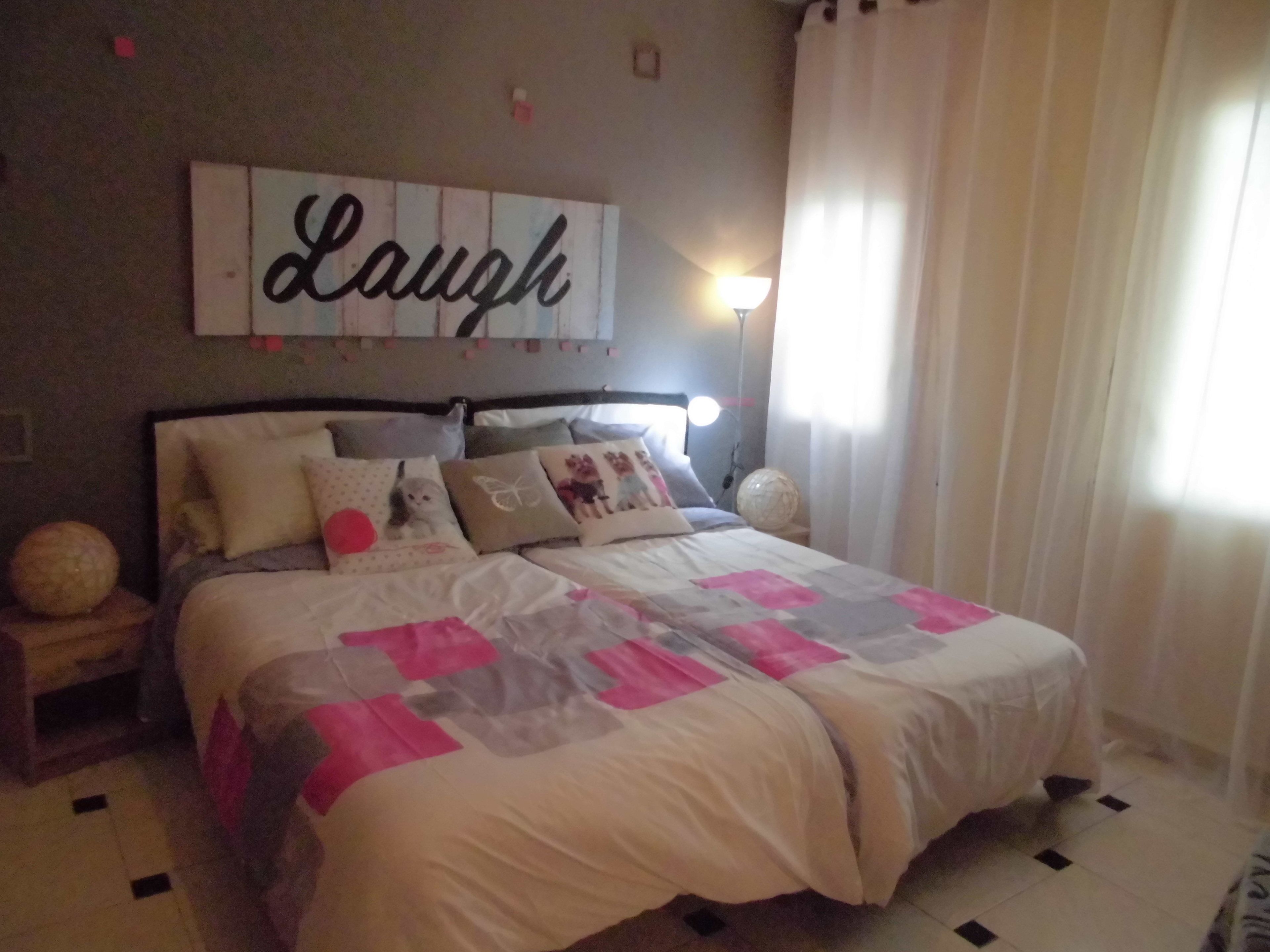 A bedroom with a bed, pillows, a wall sign, a nightstand, and a lamp.