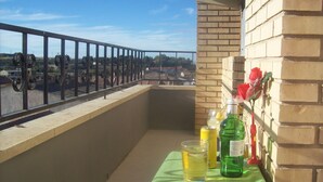 Un balcón con una mesa dispuesta para dos personas, con mantel verde, un vaso de agua, una lata de refresco y una botella de líquido verde. Se ve un muro de ladrillo y una barandilla al fondo.