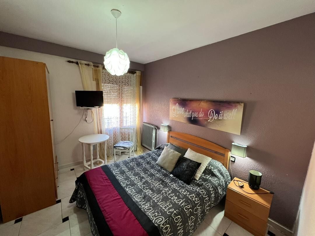 A bedroom with a bed, bedside tables, a TV, a small table, and a window with curtains.