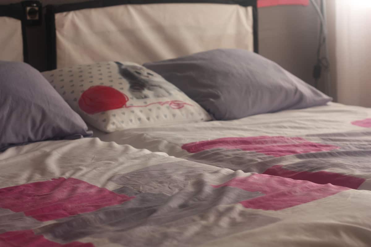 Een bed met een wit en roze sprei, een kussen met een rood ontwerp, en een hoofdbord.