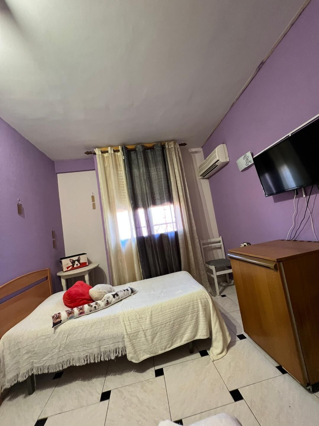 A bedroom with a bed, a desk, a chair, a TV, and an air conditioning unit.