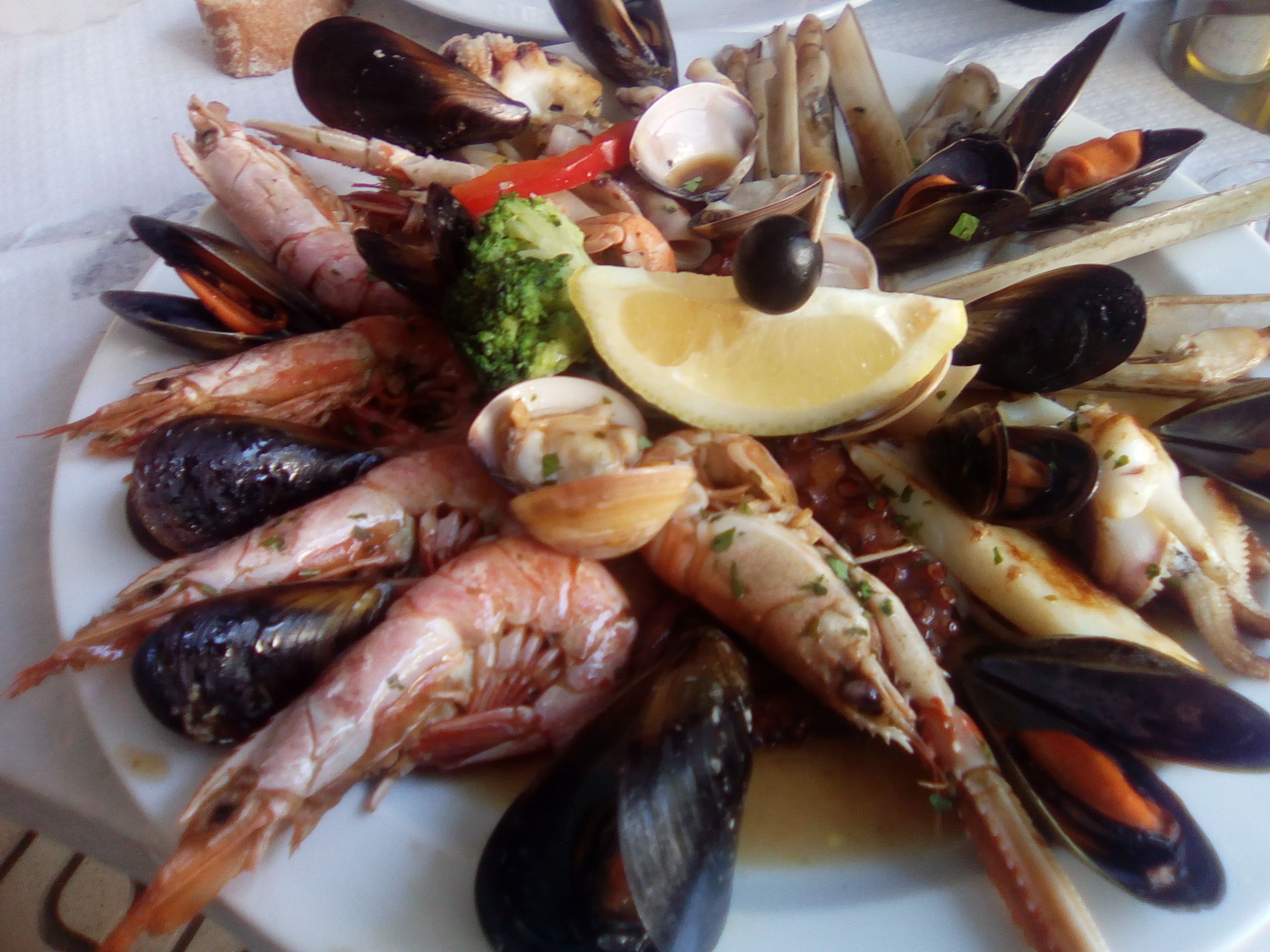 Una bandeja de mariscos con diversos moluscos, como camarones, mejillones y almejas, adornada con una cuña de limón y una aceituna negra.