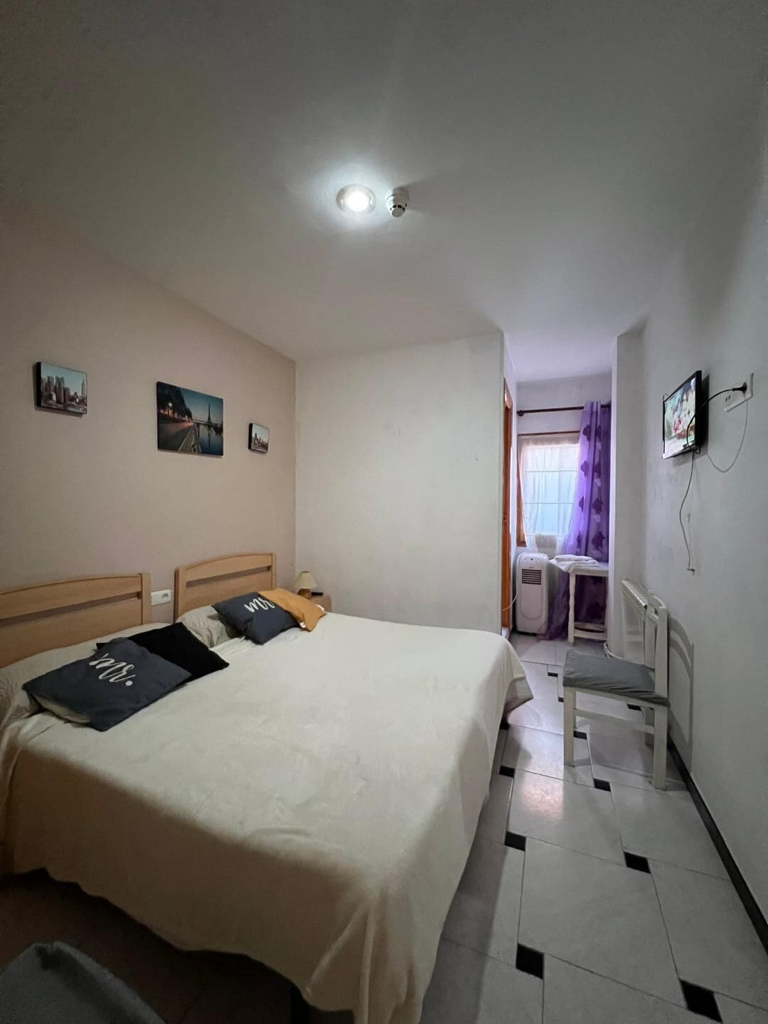 A hotel room with a bed, a nightstand, a wall-mounted TV, and a window with curtains.
