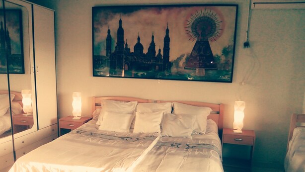 Una cama bien hecha con sábanas blancas en una habitación de hotel, un cuadro grande de un paisaje urbano sobre la cama y dos lámparas de noche sobre mesitas de noche.