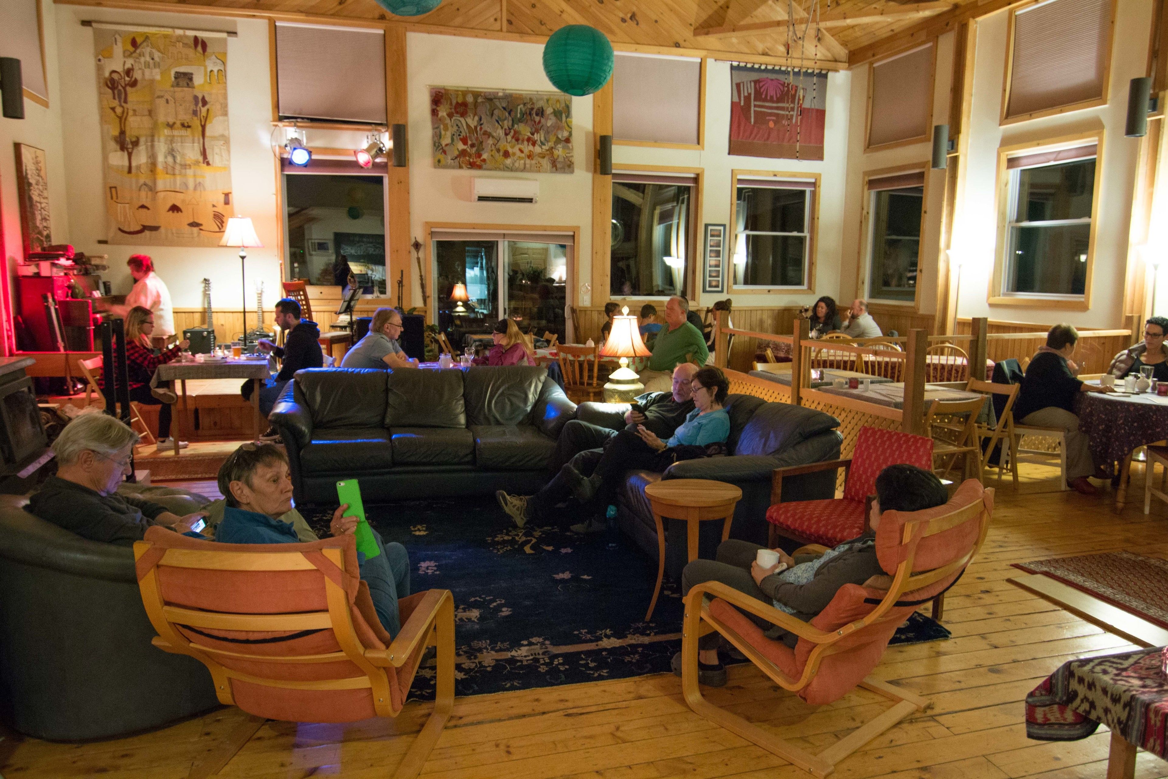 A cozy indoor space with wooden furniture, including sofas and chairs, arranged around a central area with a rug. There are tables and chairs for dining, and the room has large windows and a high ceiling.