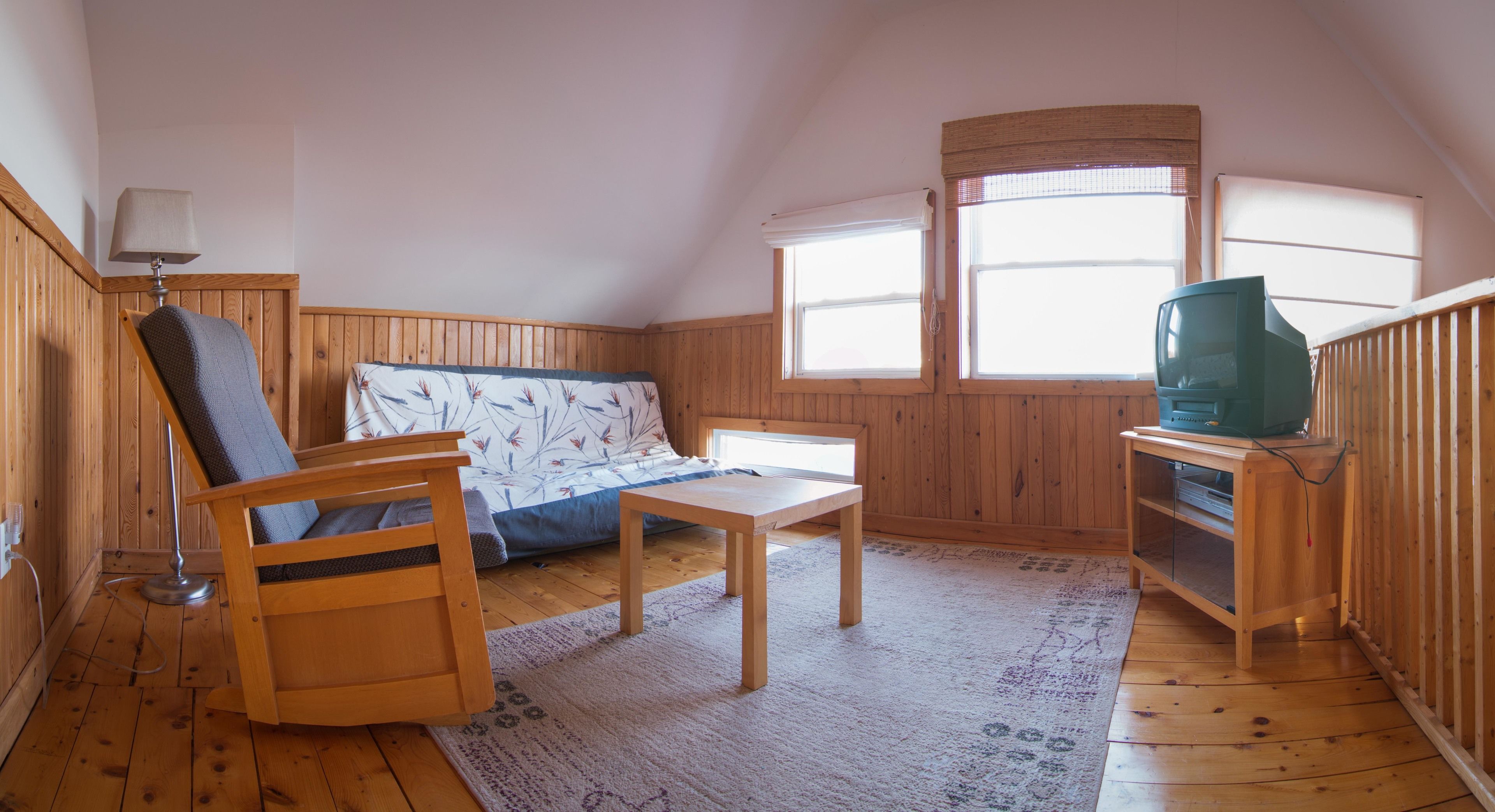 Una habitación acogedora con suelo de madera, un sofá estampado con flores, una mesa de madera, un televisor sobre un mueble de madera y una ventana con persianas.