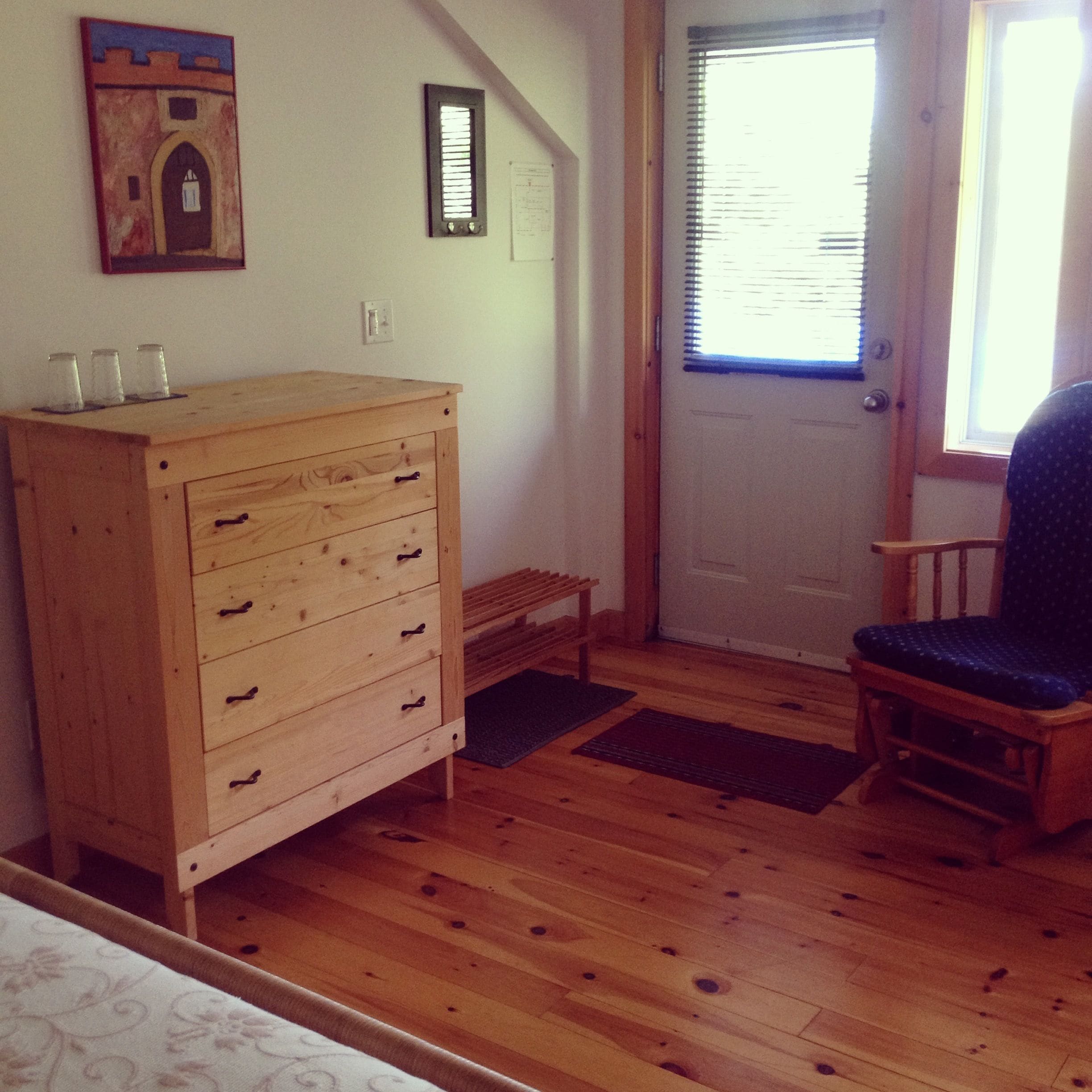 A room with a wooden dresser, a chair, a door with blinds, and a small bench.
