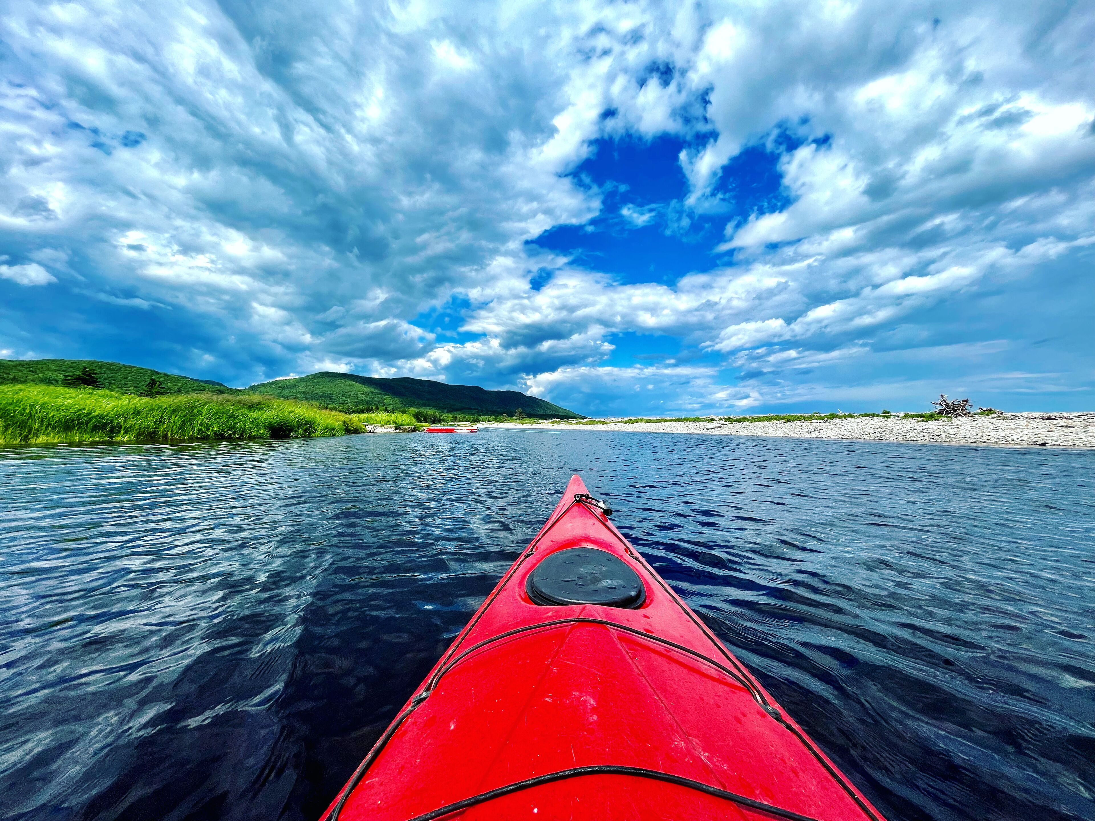 A red kayak on calm water with a view of a riverbank and distant hills under a partly cloudy sky.