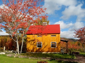 Uma cabana de madeira com telhado vermelho, rodeada por folhagem outonal e um céu claro.