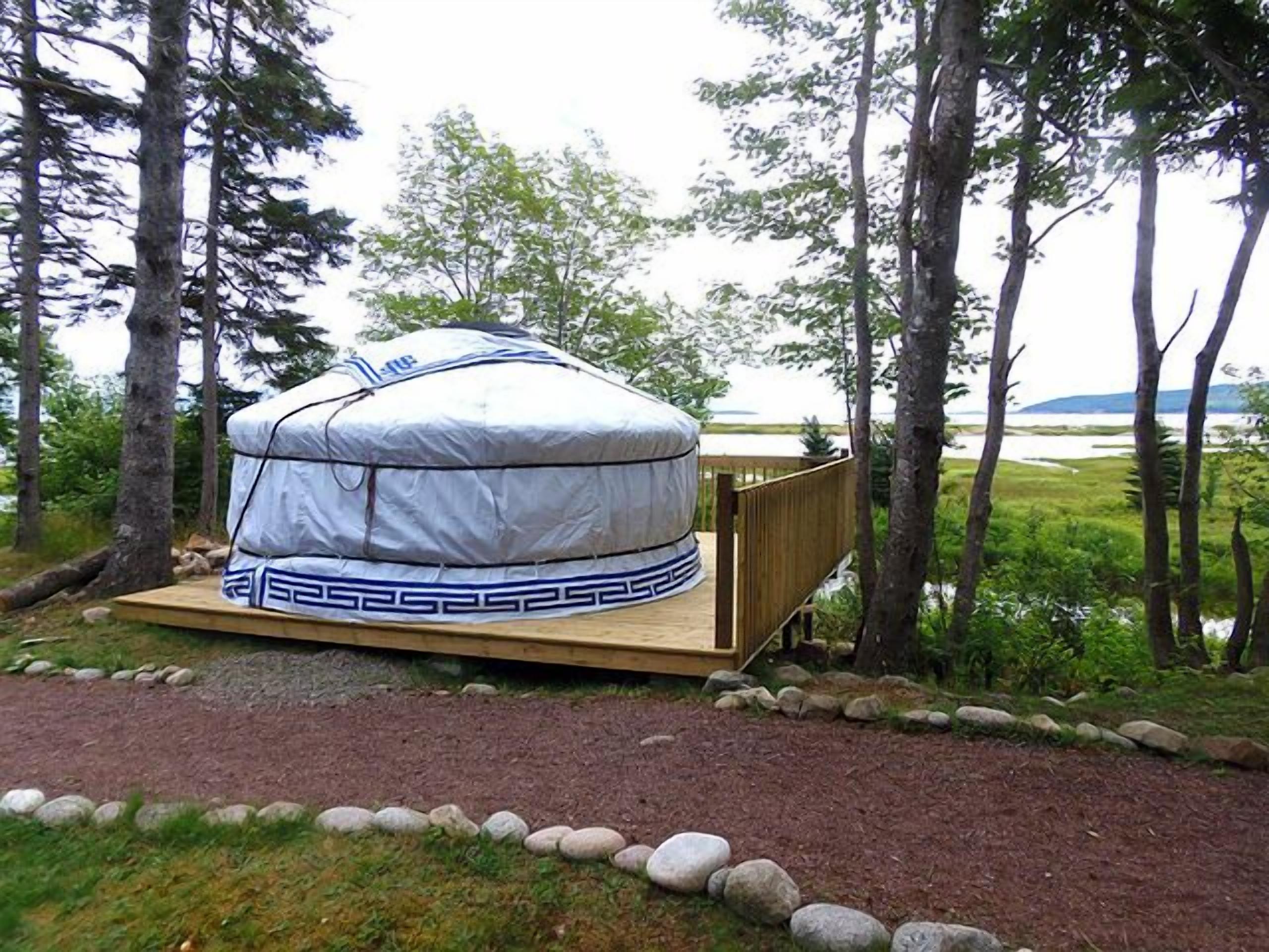 A yurt with a wooden platform, surrounded by a natural landscape with trees and a body of water in the background.
