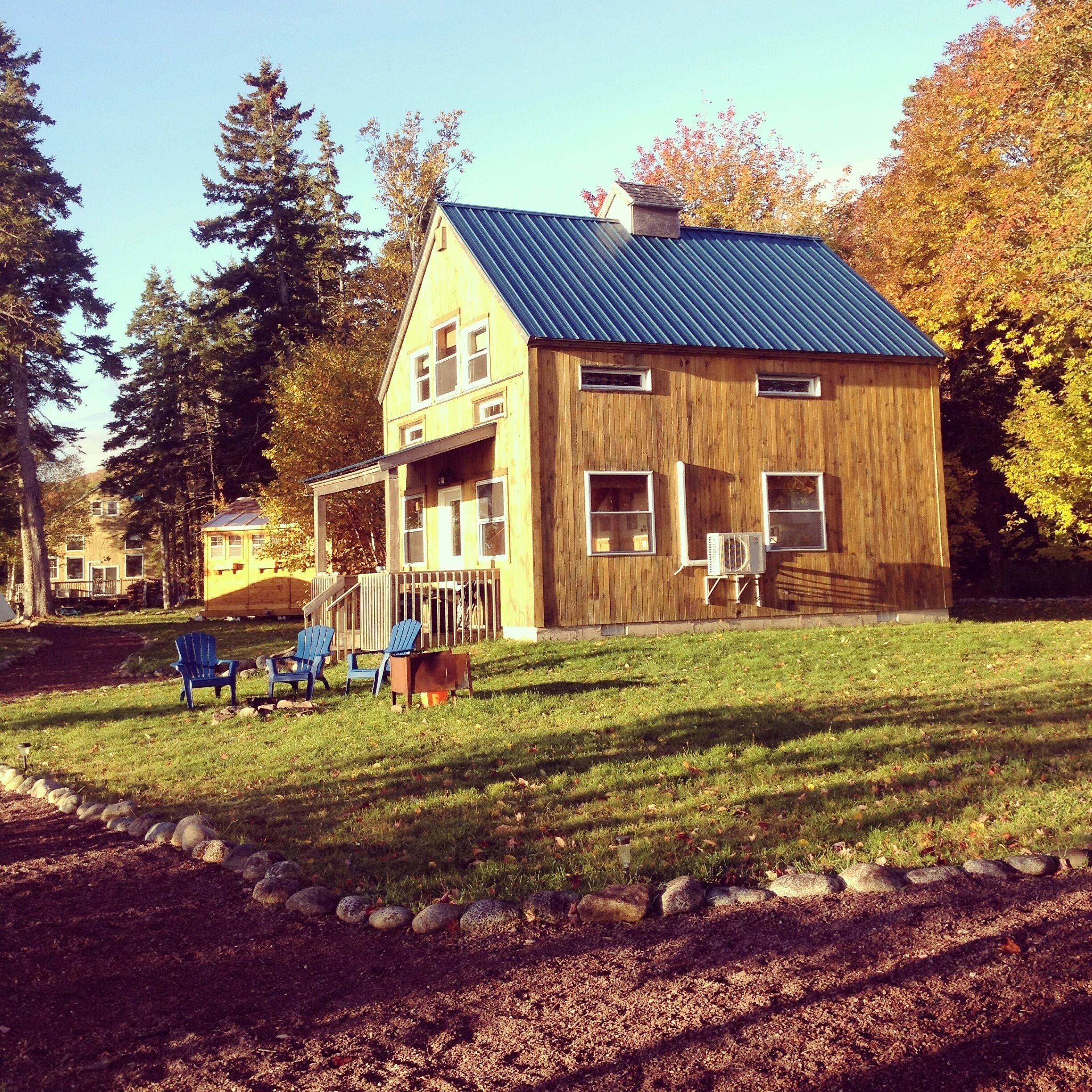 Une maison en bois avec un toit en métal, une véranda dotée de chaises Adirondack bleues et une pelouse bordée de pierres.