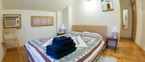 Un dormitorio con cama, una escalera de madera, aire acondicionado de pared, una ventana con persiana, una mesita de noche con lámpara y una puerta con espejo.