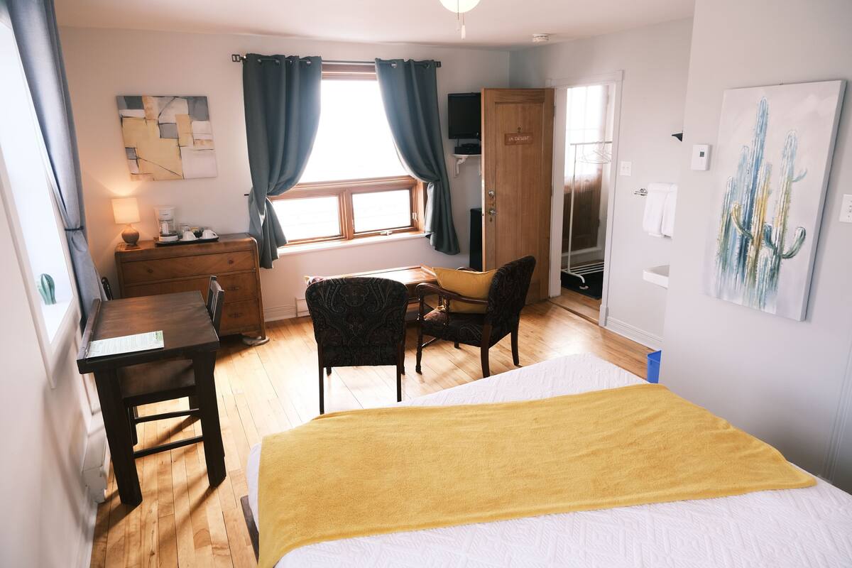 A bedroom with a bed, a desk, a chair, a dresser, a window with curtains, and a painting on the wall.
