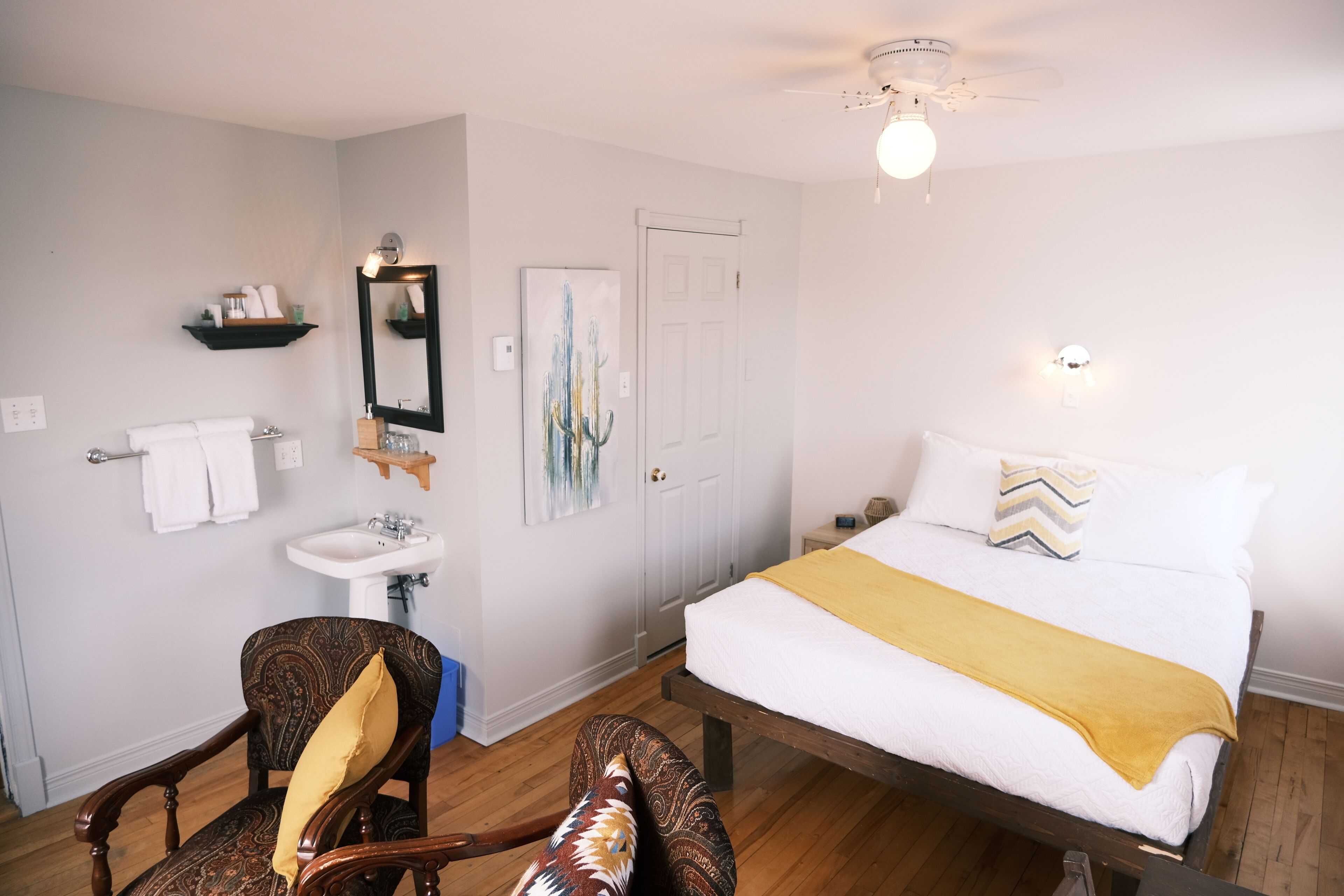 Un dormitorio con cama, una silla, mesita de noche, espejo, repisa con artículos de aseo y una puerta.