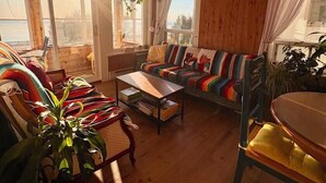 Ein gemütliches Wohnzimmer mit Holzboden, einem farbenfrohen Sofa, einem Couchtisch und einem Esstisch. Große Fenster lassen natürliches Licht herein.