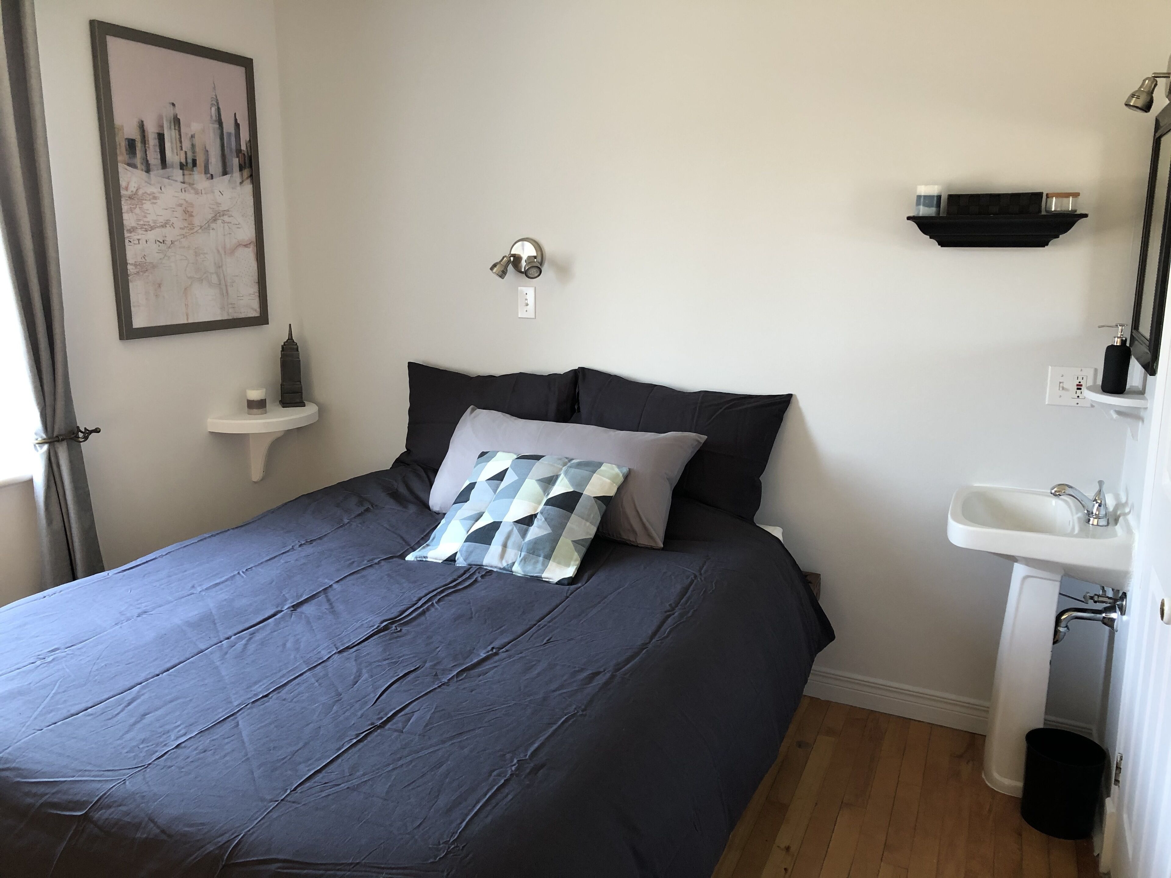 A neatly made bed with a dark blue bedspread and pillows, a small bedside table with a lamp, a wall-mounted shelf with a mirror, and a white sink with a black bucket.