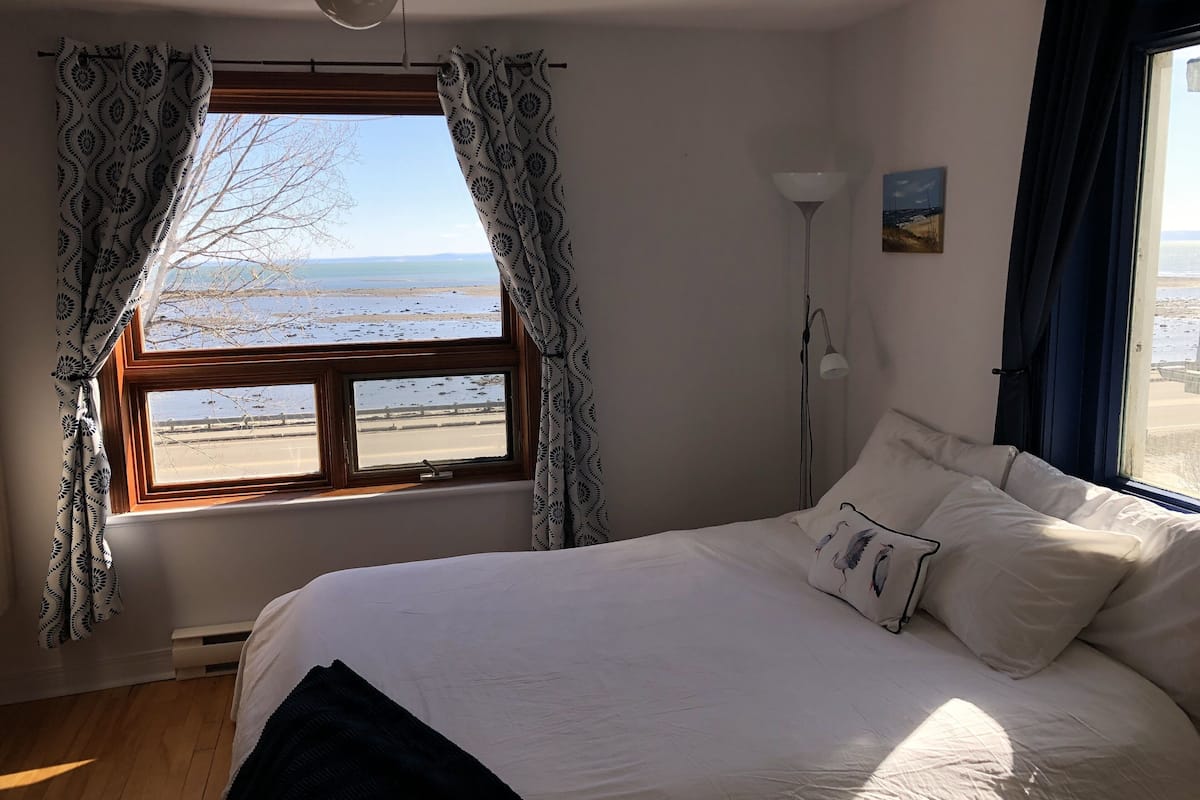 A bedroom with a large bed, a window with patterned curtains, a standing lamp, and a painting on the wall.