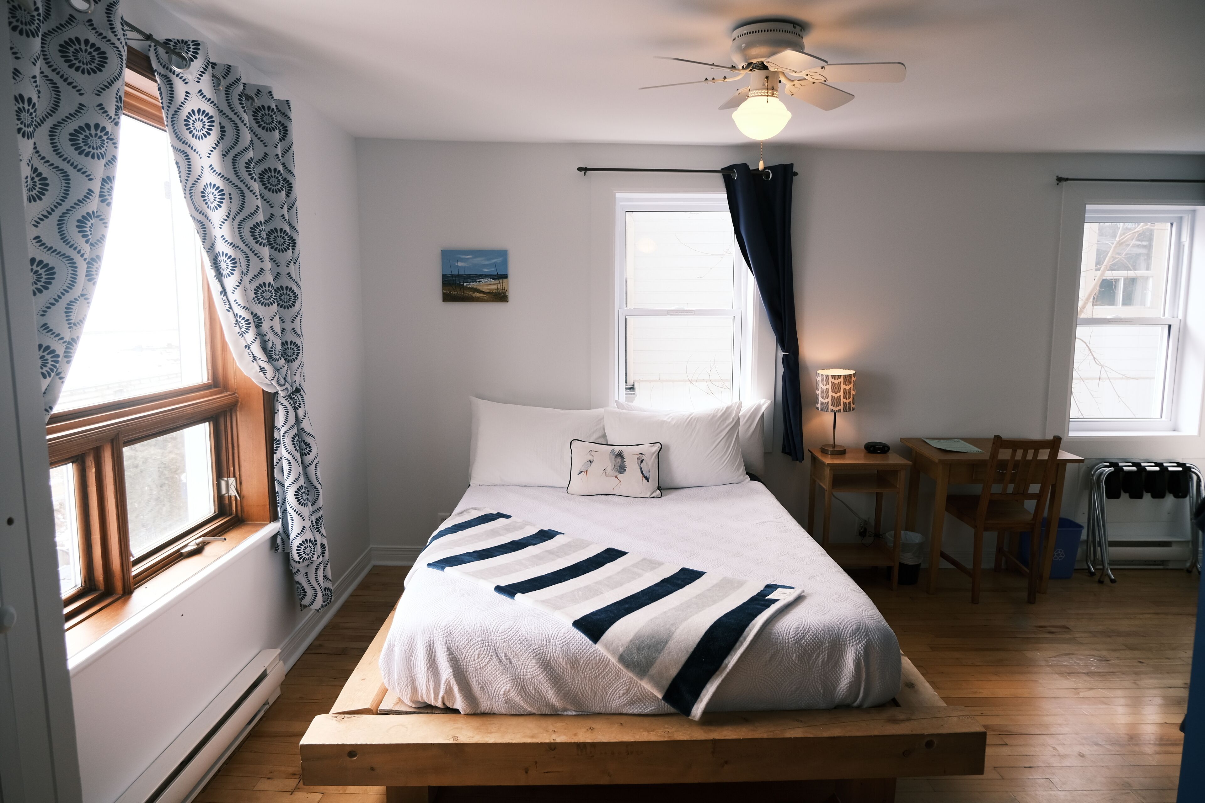 Quarto com cama, escrivaninha e cadeira. Há uma janela com cortinas, um ventilador de teto e um abajur.