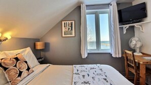 Ein Schlafzimmer mit einem Bett, einem Nachttisch mit Lampe, einem an der Wand montierten Fernseher, einem Fenster mit Vorhängen und einem kleinen Schreibtisch mit Stuhl.