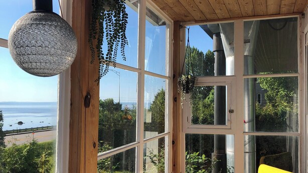 Ein raum mit Holzwänden, einer Bank und einem gelben Stuhl, große Fenster mit Blick auf das Meer und viel Grün.