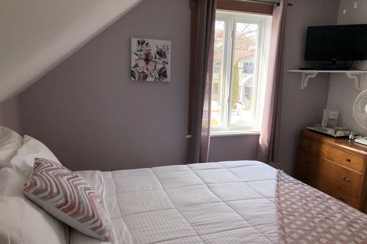A bedroom with a bed, a television on a shelf, a window with curtains, and a dresser.