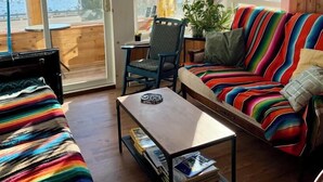 Ein gemütliches Wohnzimmer mit einem farbenfrohen, gestreiften Sofa, einem kleinen Tisch und einem Sessel. Durch große Fenster ist der Blick auf das Meer möglich.